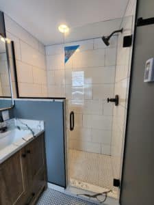 Modern bathroom with a glass-enclosed shower featuring white subway tiles on the walls and small hexagonal tiles on the floor. The vanity has a marble-patterned countertop, a wooden cabinet, and a round mirror, adding a contemporary touch.