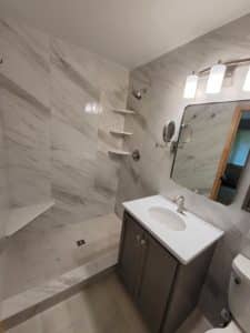 Bathroom with a sleek, modern vanity featuring a white countertop and an undermount sink, paired with a dark cabinet. Above the vanity, there's a wall-mounted mirror and a stylish light fixture. The shower area has large marble-look tiles, built-in corner shelves, and a minimalist design.