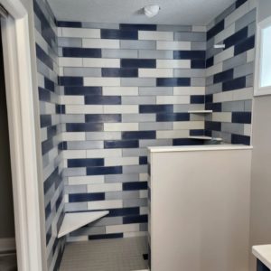 Shower area with a bold tile design featuring alternating dark blue, light blue, and white rectangular tiles arranged in a staggered pattern. The space includes built-in corner shelves and a partial wall for added privacy.