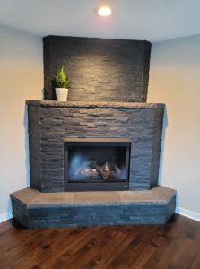 A corner fireplace with a black stacked stone surround, a tiled hearth, and a small plant on the mantel.